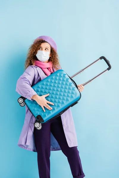 Mujer rizada en boina, bufanda, máscara médica y abrigo que sostiene el equipaje en azul - foto de stock