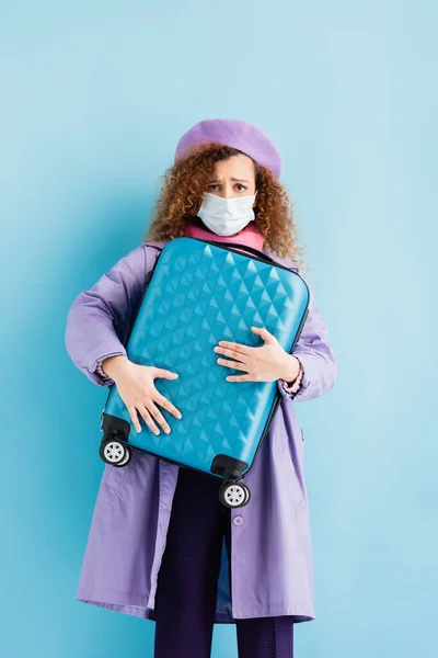 Сумна кучерява жінка в береті, шарфі, медичній масці та пальто тримає валізу на синьому — стокове фото