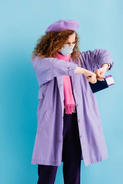 Curly woman in beret, scarf, medical mask and coat tearing passport on blue — Stock Photo