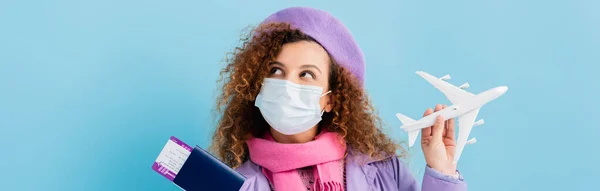 Donna riccia in berretto, sciarpa e maschera medica in possesso di passaporto e aereo giocattolo su blu, banner — Foto stock