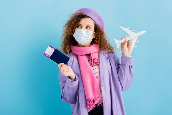 Femme bouclée en béret, écharpe, masque médical et manteau tenant passeport et avion jouet sur bleu — Photo de stock