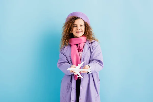 Mulher positiva em boina, cachecol e casaco segurando avião de brinquedo em azul — Fotografia de Stock