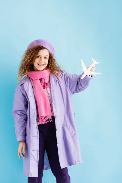 Jovem mulher positiva em boina, cachecol e casaco segurando avião de brinquedo em azul — Fotografia de Stock