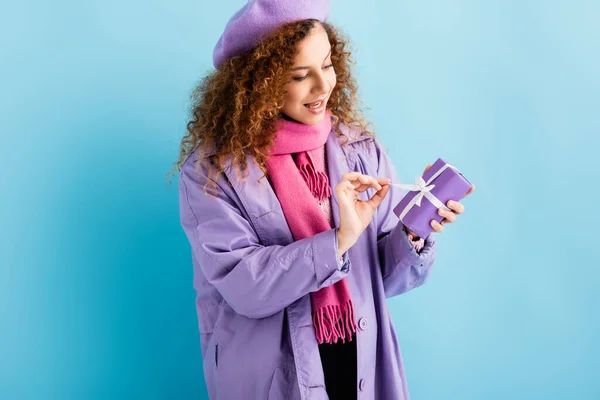 Happy woman in winter coat, beret and pink knitted scarf untying ribbon on christmas present on blue — Stock Photo