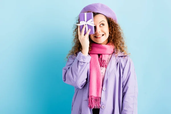 Happy woman in winter coat, beret and pink knitted scarf holding christmas present near face on blue — Stock Photo