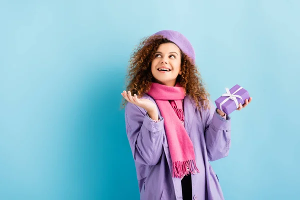 Smiling woman in winter coat, beret and pink knitted scarf holding christmas present on blue — Stock Photo