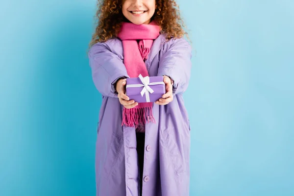 Vue recadrée de la femme souriante en manteau d'hiver et écharpe tricotée rose tenant Noël présent sur bleu — Photo de stock