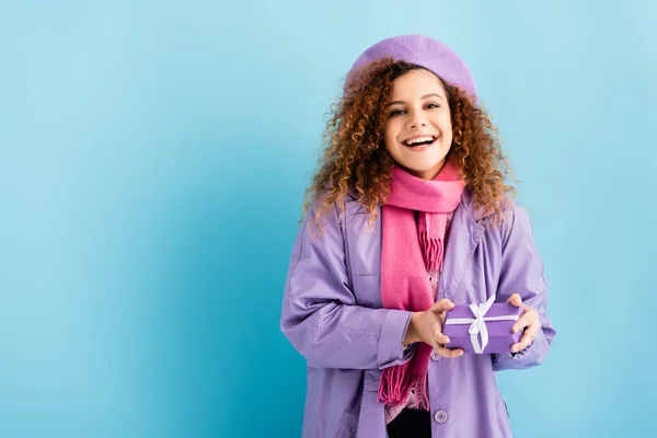Lächelnde junge Frau in Wintermantel, Baskenmütze und rosa Strickschal mit Weihnachtsgeschenk auf blauem Grund — Stockfoto