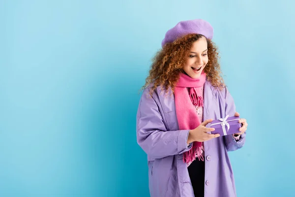 Young amazed woman in winter coat, beret and pink knitted scarf holding christmas present on blue — Stock Photo