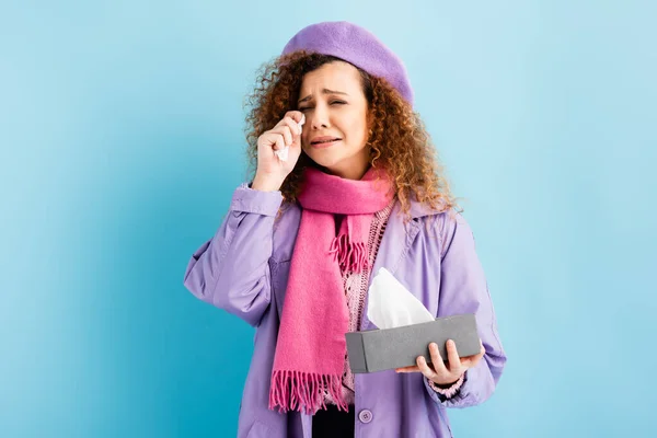 Upset young woman in winter coat, beret and pink knitted scarf holding tissue box and napkin while crying on blue — Stock Photo
