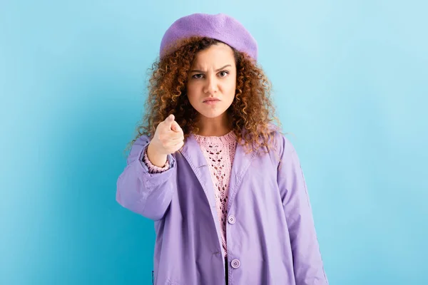 Angry woman in beret and coat pointing with finger while looking at camera on blue — Stock Photo