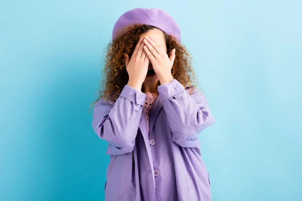 Scared and curly woman in beret covering face with hands on blue — Stock Photo