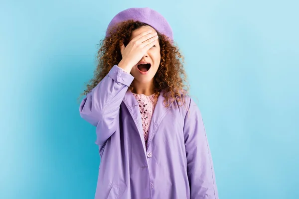 Mujer asustada y rizada en boina cubriendo la cara con las manos y gritando en azul - foto de stock