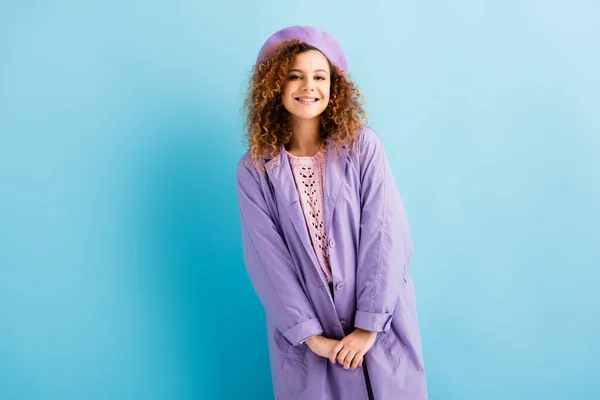 Mujer joven positiva en boina y abrigo mirando hacia otro lado en azul - foto de stock
