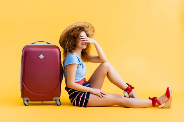 Весела і кучерява жінка прикриває обличчя, сміючись і сидячи біля валізи на жовтому — стокове фото