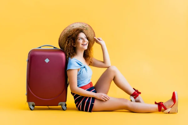 Cheerful and curly woman adjusting straw hat while laughing and sitting near suitcase on yellow — Stock Photo