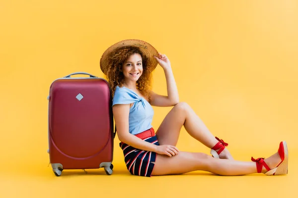 Femme heureuse et bouclée ajustant chapeau de paille tout en étant assis près de la valise sur jaune — Photo de stock