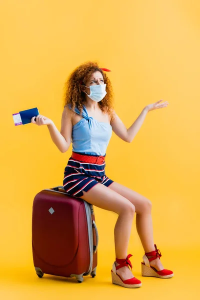 Confus jeune femme dans le masque médical tenant passeport tout en étant assis sur la valise sur jaune — Photo de stock