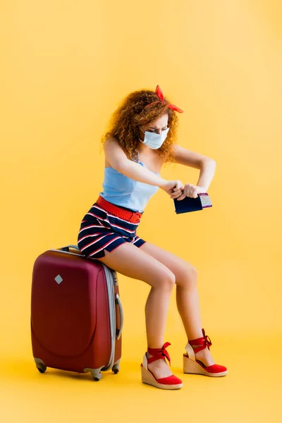 Jeune femme bouclée dans le masque médical déchirant passeport tout en étant assis sur la valise sur jaune — Photo de stock