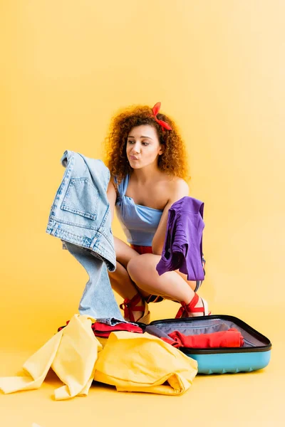 Confused woman sitting near suitcase and choosing between clothing on yellow — Stock Photo