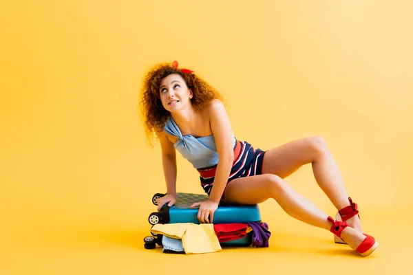 Curly young woman sitting on suitcase with clothing on yellow — Stock Photo