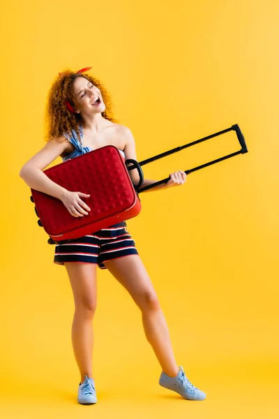 Piena lunghezza di donna felice in abito estivo e scarpe da ginnastica che tengono la valigia rossa e cantano sul giallo — Foto stock