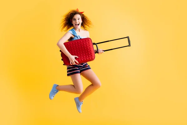 Longitud completa de la mujer feliz en traje de verano y zapatillas de deporte saltando con la maleta en amarillo - foto de stock