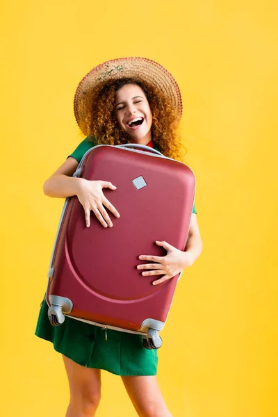 Mujer sorprendida en sombrero de paja sosteniendo equipaje rojo aislado en amarillo — Stock Photo