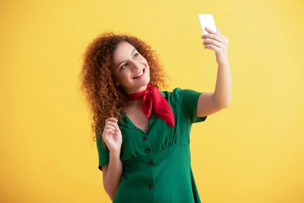 Encaracolado jovem mulher em vestido verde sorrindo ao tomar selfie no amarelo — Fotografia de Stock