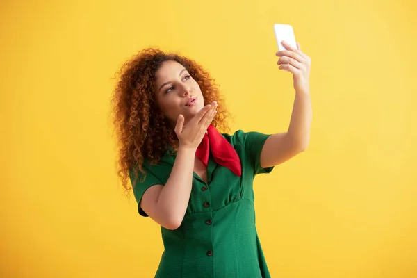 Rizado joven mujer en verde vestido enviando aire beso mientras toma selfie en amarillo - foto de stock