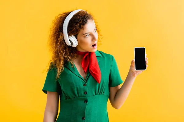 Mulher chocada em fones de ouvido sem fio olhando para smartphone com tela em branco isolada no amarelo — Fotografia de Stock