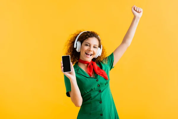 Mujer alegre en auriculares inalámbricos sosteniendo teléfono inteligente con pantalla en blanco aislado en amarillo - foto de stock