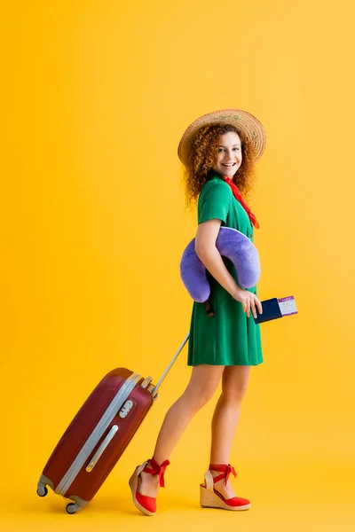 Повна довжина щасливої жінки в солом'яному капелюсі, що тримає паспорт, подушку для подорожей і ходьби з багажем на жовтому — стокове фото