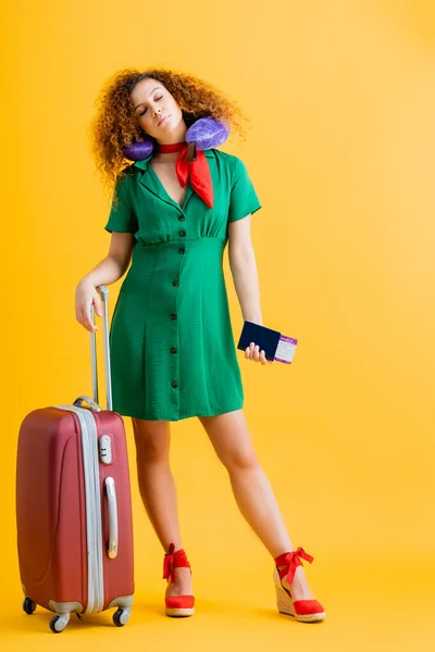 Full length of sleepy tourist in green dress with travel pillow holding passport near suitcase on yellow — Stock Photo