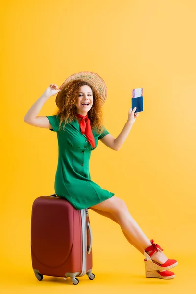 Full length of happy woman in straw hat holding passport and sitting on red luggage on yellow — Stock Photo
