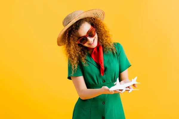 Smiling woman in straw hat, sunglasses and dress holding toy plane on yellow — Stock Photo