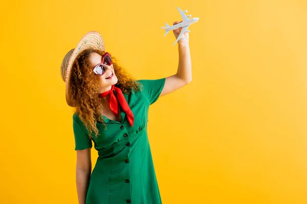 Mulher positiva em chapéu de palha, óculos de sol e vestido segurando avião de brinquedo no amarelo — Fotografia de Stock