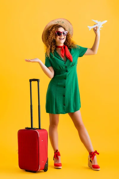 In voller Länge glückliche Frau mit Strohhut, Sonnenbrille und Kleid, die Spielzeugflugzeug in der Nähe von Gepäck auf gelb hält — Stockfoto
