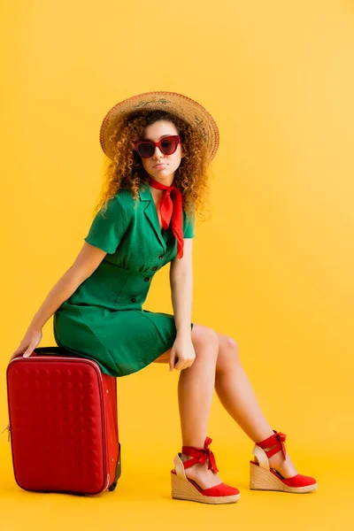 Piena lunghezza di donna triste in cappello di paglia, occhiali da sole e vestito seduto su bagagli su giallo — Foto stock