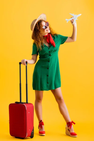 Повна довжина веселої жінки в солом'яному капелюсі, сонцезахисних окулярах і сукні, що тримає іграшковий літак біля багажу на жовтому — стокове фото