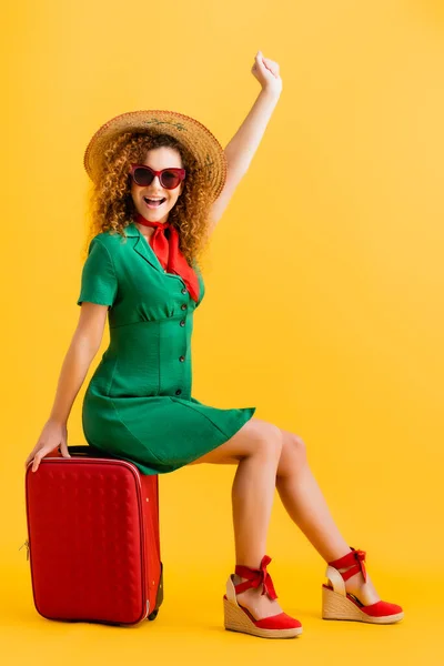 Full length of excited woman in straw hat, sunglasses and dress sitting on luggage on yellow — Stock Photo