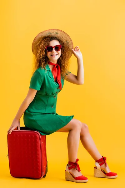 Volle Länge der fröhlichen Frau mit Strohhut, Sonnenbrille und Kleid sitzt auf Gepäck auf gelb — Stockfoto