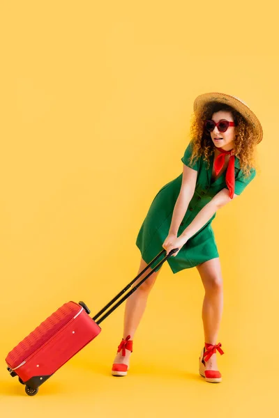 Longitud completa de la mujer en sombrero de paja, gafas de sol y vestido tirando de equipaje en amarillo - foto de stock