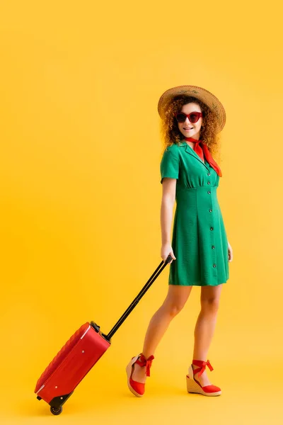 In voller Länge fröhliche Frau mit Strohhut, Sonnenbrille und Kleid mit Gepäck auf gelb — Stockfoto