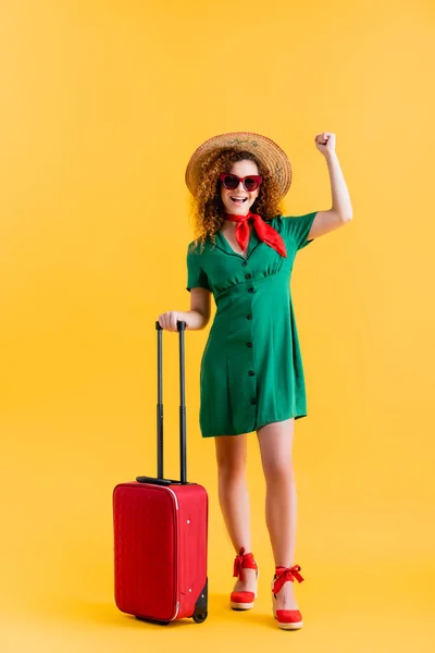 Повна довжина збудженої жінки в солом'яному капелюсі, сонцезахисних окулярах і сукні, що стоїть з багажем на жовтому — стокове фото