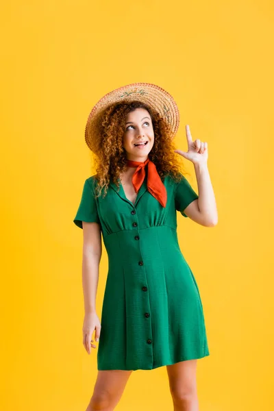 Cheerful young woman in straw hat and green dress pointing with finger and looking up isolated on yellow — Stock Photo