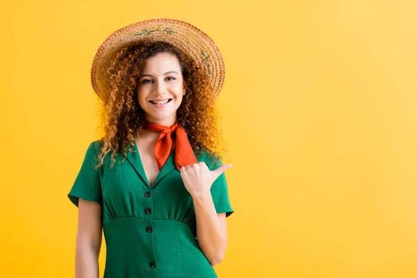 Happy young woman in straw hat and green dress pointing with thumb isolated on yellow — Stock Photo