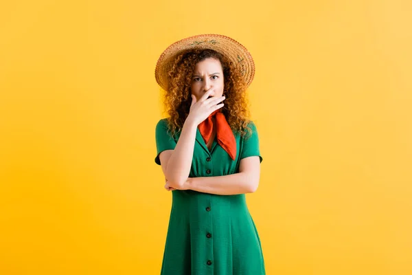Scared young woman in straw hat and green dress covering mouth on yellow — Stock Photo