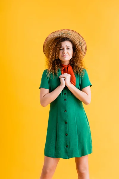 Traurige und lockige Frau mit Strohhut und grünem Kleid, die mit geballten Händen auf gelb steht — Stockfoto