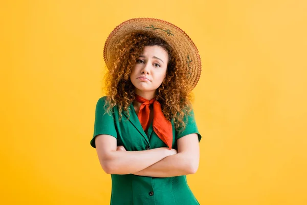 Sad and curly woman in straw hat and green dress standing with crossed arms on yellow — Stock Photo